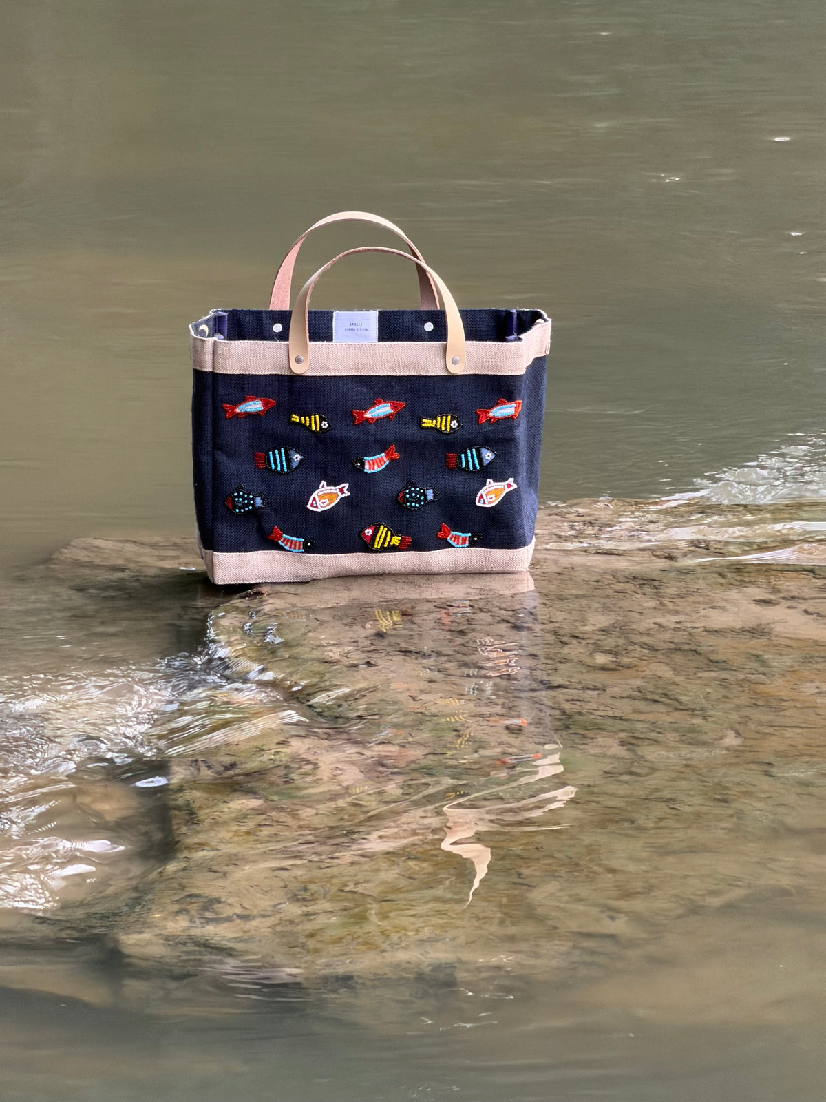 Navy Petite Market Bag with multicolored beaded fish, cream trim, and light leather handles, photographed beside a creek.