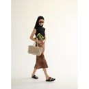 Petite Mercado in Natural — full-length view, carried in hand while walking in black slide sandals, showing the bag's scale against the body
